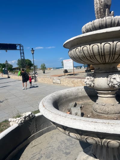 The image shows an outdoor scene on a sunny day, with a bright blue sky and scattered white clouds. The camera is tilted, giving a dramatic perspective of a large, ornate stone structure, possibly a fountain or monument, in the foreground. The stone has intricate carvings, including what appear to be lion heads and fluted patterns.
In the mid-ground, a paved pathway runs alongside the stone structure. Several people are visible on the pathway. One person in the foreground is wearing a black jacket and shorts and appears to be walking away from the camera, holding a bright green object. Another person in a red top and dark shorts is standing closer to the stone structure. In the background, more people are seen near a beach or waterfront area. Trees and lampposts are also present.
Further in the background, a body of water is visible, suggesting a coastal or lakeside setting. A sign is posted on the pathway with text that is partially visible. The overall impression is of a public space, possibly a park or promenade, in a Mediterranean or similar climate, given the clear skies and outdoor activity. The time of day appears to be midday or afternoon, judging by the bright sunlight and defined shadows.