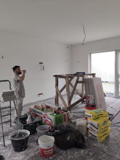 A man is taking a break from work inside a room that is under renovation. He is standing on the left side of the image, wearing a grey, paint-splattered t-shirt and pants, and is drinking from a bottle. The room is a bright, empty space with white walls. Electrical outlets are visible on one wall, and a bare lightbulb hangs from the ceiling.
Construction materials are scattered across the floor, which is covered with a plastic tarp. A wooden scaffolding structure stands in the center of the room, supporting some tools and building materials. To the right, a stack of yellow bags with the brand name "Vetonit" are piled up, along with other construction supplies. Insulation boards labeled "ТЕХНОНИКОЛЬ XPS ТЕХНОПЛЕКС" lean against the scaffolding. Several buckets, including one with "KNAUF ROTBAND PASTA" written on it, are also present. The overall scene suggests an ongoing renovation project, with the man enjoying a moment of rest amidst the building materials.