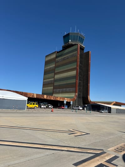 The image shows the air traffic control tower at Lleida-Alguaire Airport, located in Alguaire, Spain. The tower is a tall, modern structure with a glass-enclosed control room at the top. A large, rust-colored canopy extends from the tower's base, with the word "ALGUAIR" vertically oriented. Several vehicles, including a yellow van, are parked at the base of the tower. The ground is concrete and has painted markings. The sky is clear and blue.