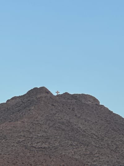A lone cross stands atop a rocky mountain peak against a clear, pale blue sky. The mountain is covered in dark, rough terrain, with the cross positioned slightly to the right of the center of the frame, its silhouette visible against the sky. The image appears to be a photograph taken from a distance, capturing the vastness of the landscape and the isolated prominence of the cross. The location is identified as Sunland Park, United States of America.