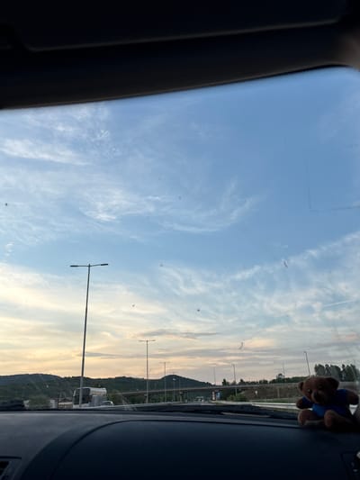 The image captures a view from inside a vehicle, looking through the windshield at a highway landscape under a cloudy sky at dusk. In the foreground, the dark dashboard of the car is visible, with a small, brown teddy bear wearing a blue shirt perched on the right side. Beyond the windshield, a highway stretches into the distance, lined with lampposts. On the left, a hill covered in trees slopes down towards the road. A large, white bus is stopped on the highway, and other vehicles are visible. The sky is a mix of blue and orange hues, indicating the setting sun, with wispy clouds scattered across it. The overall scene suggests a journey on a highway near Mursalevo, Bulgaria, during the evening.