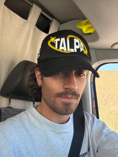 A man is seated in a vehicle, wearing a black baseball cap with yellow lettering "TALPA" and a star emblem. He has a light beard, brown hair, and is wearing a light gray long-sleeved shirt and a seatbelt. He appears to be resting his head on a black headrest. A yellow cloth is partially visible in a compartment near the top left of the image. Through the window, a landscape is visible. The location is Montgenèvre, France.