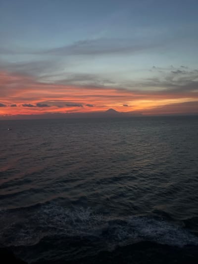 The image depicts a panoramic view of the ocean at dusk, specifically from Desa Batu Layar Barat, Indonesia. The scene is dominated by a vibrant sunset sky and a vast expanse of water.
The upper portion of the frame features a gradient sky, transitioning from intense orange and red hues at the horizon to lighter orange, then pale blue, and finally a deeper blue-grey towards the top. Stratiform clouds are visible as horizontal streaks within the orange and red bands, with sparser, darker clouds present in the blue-grey sky above.
At the horizon line, a large, dark, conical mountain, likely a volcano (possibly Mount Agung), is silhouetted against the brightly colored sky, positioned slightly to the right of the image center. Clouds obscure its lower slopes and summit.
The lower portion of the image shows the dark, undulating surface of the ocean. The water appears deep blue-grey, reflecting the diminished ambient light. Visible wave patterns create textures across the surface, with some white foam present closer to the foreground. Two small, faint white lights, indicative of distant boats, are visible on the water: one near the far left edge and another very small light near the far right. No people or overt structures are visible in the foreground or midground.