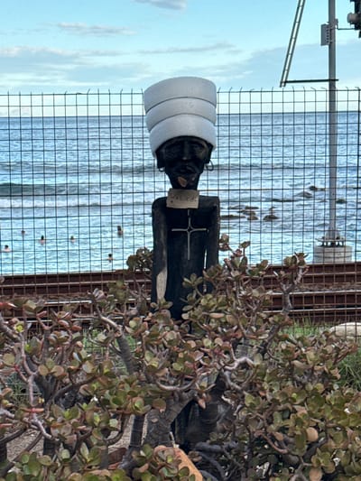 A statue stands behind a wire fence with the ocean visible in the background. The statue is dark and has a white cylindrical object on its head. A piece of paper with the word "ALLIEDS" written on it is attached to the statue's chest. A plant with small, round leaves is to the right of the statue. A metal structure is visible to the left. The location is Muizenberg, South Africa.