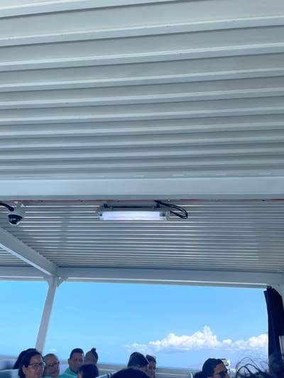 The image depicts a group of people looking out from under a corrugated metal structure, possibly a roof or awning. A fluorescent light fixture is mounted on the metal structure along with a surveillance camera. The sky is visible through an opening in the structure and contains a scattering of clouds. No identifying landmarks indicating the location in Unknown, Venezuela are visible.