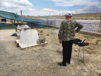 In Egizbulak, Uzbekistan, a man wearing a camouflage long-sleeved shirt, black pants, black sandals, and a white baseball cap stands with his back to the camera, facing away from two traditional tandoor ovens. The tandoor ovens, constructed from white plaster, are situated on concrete bases. Smoke emanates from the top of the oven on the left. Nearby, there are two metal buckets and a piece of fabric. To the right of the man, a small, rusty portable grill with cooking utensils and a plastic bottle is placed on the ground. The scene is set in a dry, dusty outdoor environment with sparse vegetation. In the background, a green mesh structure is visible, supported by wooden poles, and a metal fence encloses a white banner. The sky is partly cloudy with a vast expanse of rolling hills in the distance.