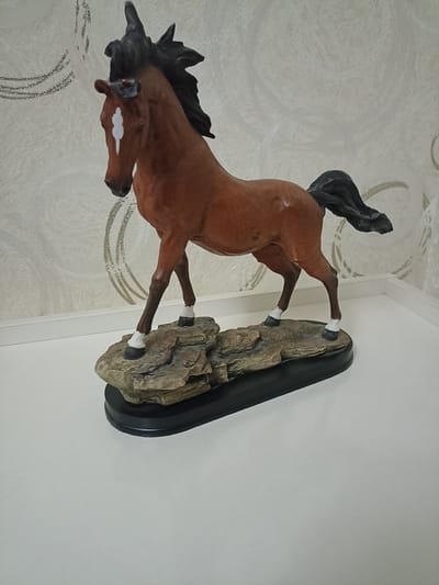 A figurine of a brown horse with a black mane and tail stands on a rocky base, which is mounted on a black oval plaque. The horse has a white marking on its forehead and white socks on its legs. The background features textured wallpaper with a swirling pattern and some shimmering silver embellishments. The figurine is placed on a white surface, likely a shelf or table. The location provided, Rabatak, Uzbekistan, is not visually discernible from the image itself.