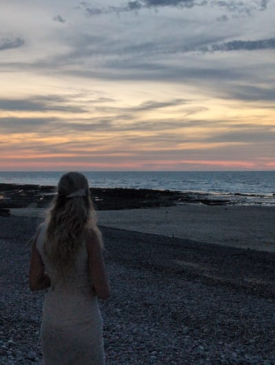 A woman with blonde, wavy hair, wearing a light-colored dress and a light-colored headband, stands facing away from the camera, looking out at the sea. Her back is to the viewer, and her hair falls past her shoulders. She is standing on a beach composed of small, dark pebbles. The sea is calm, with gentle waves, and stretches to the horizon. The sky above is a mix of gray and pale yellow clouds, with hints of pink and orange, suggesting either sunrise or sunset. In the distance, dark, rocky formations are visible along the coastline. The overall scene is a serene depiction of a coastal landscape, potentially in the city of Veules-les-Roses, France, given the context.