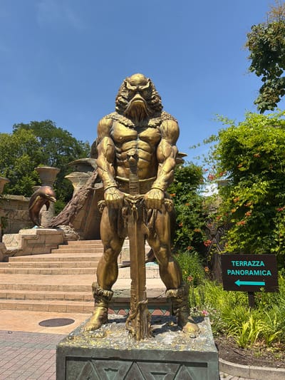 This media file showcases a large, golden statue of a muscular, mythological-looking figure standing on a stone staircase. The statue is oriented vertically in the frame, with its head near the top and feet towards the bottom. The figure appears to be wearing ornate armor and has a formidable, stern expression. To the right of the statue, a dark sign with turquoise text points to the left, indicating "TERRAZZA PANORAMICA". The background is filled with lush green foliage and a clear blue sky, suggesting a bright, sunny day. The setting appears to be an outdoor public space, possibly a park or attraction, given the signage and monumental statue. The stonework of the stairs and the base of the statue show some weathering and decorative patterns. The overall impression is one of grandeur and perhaps a hint of ancient mythology or fantasy.
