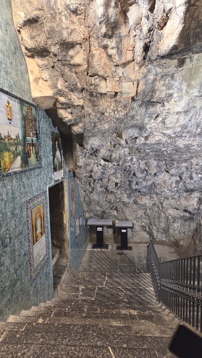 The image shows the interior of a cave-like structure in Altura, Spain. The walls are primarily rough-hewn rock. The floor is tiled with green and blue tiles and features several inset images depicting religious and historical scenes. There are openings in the floor, possibly leading to lower levels of the cave. At the top, there's a dark-colored railing. A black, rectangular object that appears to be a light fixture is mounted on the wall. The image is shot from a slightly low angle.