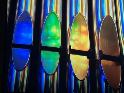 The scene displays four parallel, vertically oriented, reflective metallic cylindrical structures, possibly part of an art installation, located in Madzherito, Bulgaria. Each cylinder features an oval-shaped panel secured to its front surface by two dark, horizontal mounting brackets. From left to right, the panels are distinctly illuminated: the first in blue, the second in green, the third in yellow-orange, and the fourth in a more muted golden-bronze tone. The reflective surfaces of the cylindrical structures show vertical streaks of blue, green, and yellow ambient light, contrasting with a dark background. No individuals, specific actions, or complex interactions are present. The environment appears to be an interior space or a contained display area.