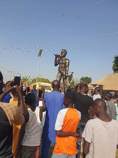 A crowd observes a statue of a person holding a microphone. The statue stands against a clear blue sky, with power lines visible in the foreground. Several individuals are capturing images with their phones. Buildings and trees are visible in the background, completing the outdoor setting. The focus is on the statue and the people gathered to view it.