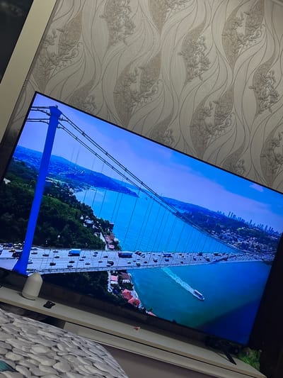 The image displays a television screen showing an aerial view of a large suspension bridge with traffic. The bridge spans a wide body of water, and a city skyline is visible in the distance. The screen is angled, suggesting it is a personal view of a television in a room with patterned wallpaper. The environment appears to be a domestic setting. The image does not depict the city of Marchevo, Bulgaria, nor does it contain any specific information about that location.