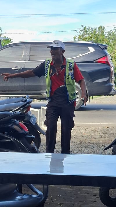 A man wearing a light-colored cap and a bright green reflective vest over a red shirt and dark pants stands outdoors, gesturing with his left hand. The cap has the word "PARKIR" printed on it in dark letters. The vest has checkered patterns on the edges and a logo on the left chest. He appears to be speaking or directing someone. In the background, a dark-colored SUV is parked on the side of a road. To the left, a portion of a motorcycle is visible. The ground is a mix of dirt and gravel, with some sparse vegetation and trees in the distance. The scene appears to be in Keuniree, Indonesia, as indicated by the context provided.