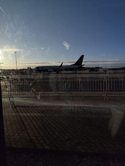 The image depicts an outdoor airfield scene at Frankfurt-Hahn Airport (HHN) in Lautzenhausen, Germany, viewed through a window, likely in the late morning or early afternoon. The foreground shows a dark, textured paved surface, possibly cobblestones, with prominent long shadows from a series of silver-colored temporary metal barrier fences that span the lower half of the frame. Reflections and subtle smudges are visible on the glass surface.
In the midground, a white and blue Ryanair Boeing 737 aircraft (registration EI-CBO) is parked on the tarmac, oriented to the right. Mobile passenger stairs are positioned at both the forward and aft passenger doors of the aircraft. Several ground crew personnel are visible around the aircraft, engaged with ground support equipment, including baggage carts. A large fuel tanker truck is parked behind the aircraft's tail section. A tall lamp post stands to the left of the aircraft's nose.
The background features a clear blue sky with scattered white clouds, particularly near the horizon and in the upper left where a bright sun flare is prominent. A faint aircraft contrail is visible in the upper left sky. A distant line of trees marks the horizon beyond the tarmac. No direct interactions between individuals are discernible; personnel appear to be conducting routine airport servicing operations.