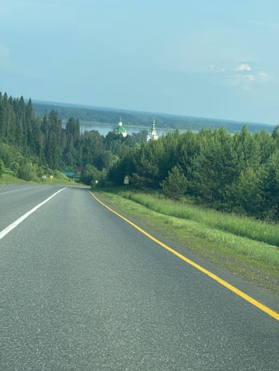 The image captures a view from a paved road, likely in or near the village of Nechkino, Russia, given the presence of a distinctive church with green domes and white spires in the background. The road has a white dividing line on the left and a solid yellow line on the right. Lush green trees and vegetation line the sides of the road, with a dense forest on the left and a mix of trees and grassy areas on the right. In the distance, a body of water, possibly a river or lake, is visible, with a village or settlement nestled amongst the trees on its banks. The sky is a pale blue with a few wispy clouds. Several road signs are present, including one with a speed limit of 50 indicated. There are no people or animals visible in the scene. The overall impression is one of a scenic rural landscape on a clear day.