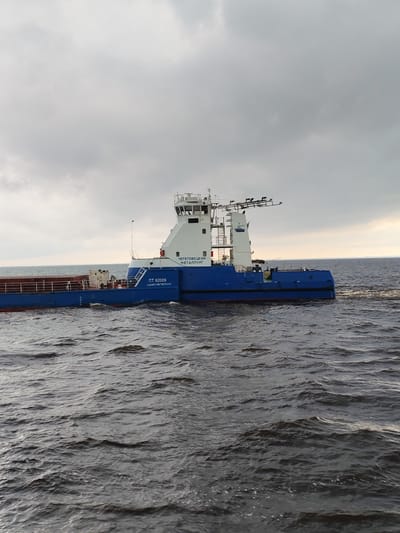 A large blue and white cargo ship is moving through dark, choppy water under an overcast sky. The ship has "ЧЕРЕПОВЕЦКИЙ МЕТАЛЛУРГ" written in Cyrillic on its side, along with "ПТ 82009" and "САНКТ-ПЕТЕРБУРГ". The ship is carrying a load of metal containers. The water appears to be a river or a lake, and the sky suggests a cloudy or stormy day. The location is identified as Novaya Svobodka, Russia.