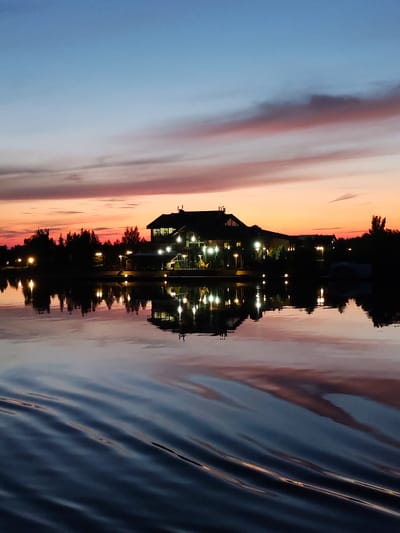 A twilight scene captures a silhouetted building complex with illuminated windows and exterior lights, situated on the shore of a body of water. The building appears to be a resort or hotel, possibly part of the Konakovo resort area in Tver Oblast, Russia, given the architectural style and waterfront setting common to the region. The sky displays a gradient of colors, transitioning from a soft blue at the top to hues of pink and orange near the horizon, indicating either sunrise or sunset. Tall, dark trees line the shore, also silhouetted against the sky. The water's surface is calm, reflecting the building's lights and the colorful sky, with gentle ripples suggesting a slight disturbance, possibly from a boat or wind. There are no visible people in the image. The overall atmosphere is serene and tranquil, characterized by the soft, diffused lighting and the peaceful reflections on the water.