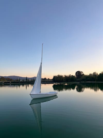 A tranquil scene features a sailboat reflected on a calm lake. The boat has white sails and its mast points towards a clear, pale blue sky transitioning to a soft, warm sunset hue. Along the lake's edge, trees create a dense border, their reflections mirrored in the still water. In the distance, subtle hints of land can be seen. The composition is serene, emphasizing the peaceful stillness of the water and the gentle light.