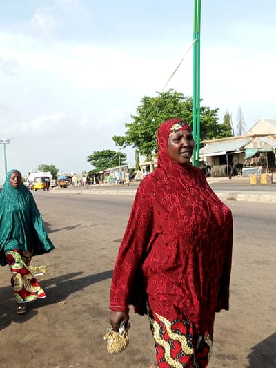 In Gadao, Nigeria, two women are captured walking along a paved road. The woman in the foreground, facing the right, is wearing a red hijab adorned with floral patterns and a matching patterned dress. She carries a small, textured pouch in her right hand. To her left, further down the road, a second woman is visible, wearing a green hijab and a colorful patterned skirt. In the background, a green pole with a wire extends upwards, and several structures including what appears to be a market with stalls, a yellow auto-rickshaw, and a large white tanker truck are present. The sky is mostly clear with some scattered clouds, and the overall environment suggests a daytime scene in a bustling urban or semi-urban area.