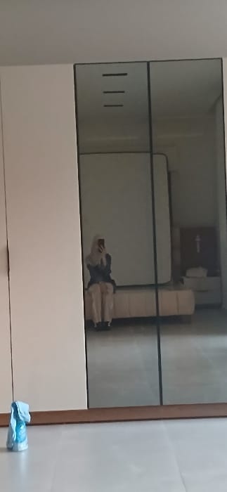 The scene depicts an indoor room in Ashrafia, Syria, focused on a large wardrobe composed of opaque white panels on the left and two mirrored panels on the right. The floor consists of light-colored, large square tiles, with a brown trim running along the base of the wardrobe. In the lower-left foreground, a light blue object, resembling a spray bottle or crumpled plastic, rests on the floor.

The mirrored section of the wardrobe reflects the interior of the room. A single individual is reflected, seated centrally on a light-colored upholstered bench or ottoman. This person is wearing a light-colored head covering (hijab), a dark long-sleeved top or jacket, and light-colored pants. They are holding a dark smartphone with both hands, angled towards the mirror, indicating they are capturing their own reflection.

Behind the reflected individual, a large, unmade light-colored mattress stands upright, leaning against a wall. To the right of the mattress, a small bedside table or chest of drawers is visible. On its surface, a white folded item and another unidentifiable object are placed. A brown surface above the bedside table prominently features a white upward-pointing arrow symbol. Near the top edge of the mirrored reflection, three short, dark horizontal lines are visible, possibly reflecting a light fixture or design element on the room's ceiling.