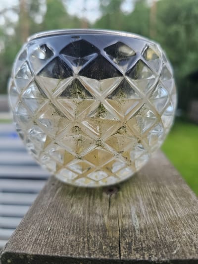 An outdoor, close-up, eye-level shot showcases a textured glass orb with a diamond pattern, resting on a weathered wooden railing. The orb, which appears to be part of a solar garden light, contains a yellow liquid and has some green discoloration on its surface. The background is softly blurred, revealing a green grassy area and trees, indicative of a natural outdoor environment. The lighting suggests it is daytime. The scene is devoid of any human presence or activity. The location is identified as Mäntsälä, Finland.