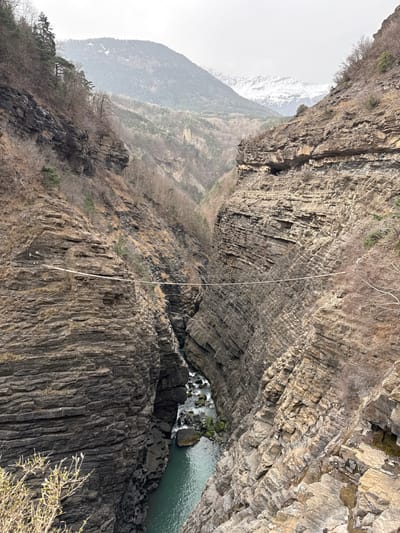 The image depicts a deep, narrow river gorge situated in a mountainous region near Corps, France. The gorge features steep, layered rock walls of dark brown and gray strata, indicative of sedimentary rock formations. A winding river, displaying sections of turquoise-green still water and areas of white rapids over submerged rocks, flows at the bottom of the chasm. The river narrows significantly further into the gorge.
A thin, light-colored line, consistent with a tightrope or slackline, is stretched horizontally across the gorge, traversing from the mid-level of the left rock face to a similar height on the right rock face. This line is secured to the rock walls, indicating human activity or preparation for such.
Sparse vegetation, consisting primarily of leafless deciduous trees and some scattered evergreen conifers, clings to the less precipitous slopes and ledges of the gorge walls. In the background, beyond the immediate gorge, multiple layers of mountains rise, covered with forests exhibiting winter dormancy. The most distant peaks in the background are partially covered with snow, under an overcast, pale grey sky. No individuals are visible in the scene.