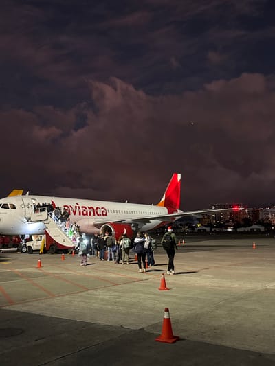 Passengers disembark an Avianca aircraft at night via a mobile stairway. Orange cones line the tarmac near the plane. The fuselage displays the Avianca logo. A person stands near the tail of the plane. In the background, other vehicles and lights are visible, indicating an airport setting. The sky is dark with clouds, possibly dusk or dawn.