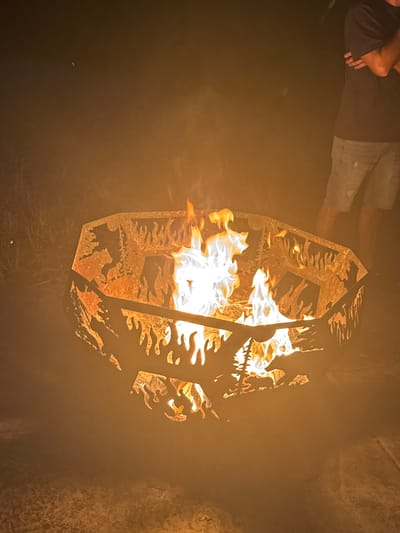 At night, a fire burns in a circular metal fire pit with cut-out designs of trees and animals. The fire pit is on a paved surface. A person's legs and torso are visible on the right side of the frame, wearing a dark t-shirt and denim shorts. The fire is bright white and yellow, with flames reaching upwards. The background is dark and out of focus, with some visible grass and brush.