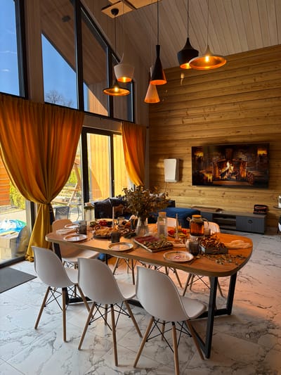 The image shows a dining area in a wooden-walled home in Marusino, Russia. The room is bright with light entering through large windows covered with golden drapes. A long, oval wooden table is centrally located, laden with food, drinks, and decorative foliage. Five white plastic chairs with wooden legs are arranged around the table. The floor is covered in light marble tiles with dark grey veining. The ceiling and walls are clad in light brown wood planks. Several modern pendant lights with varying shapes and colors (gold, black, and white) hang from the ceiling. On the wall opposite the windows is a painting of a fireplace above a white box, and a grey cabinet is to the right of the painting. The scene appears to depict a meal setting within a modern wooden home interior.