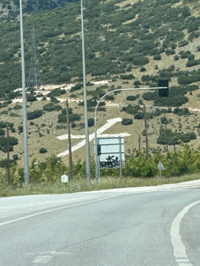 The image depicts a hillside in Prosotsani Municipal Unit, Greece, marked with a large white "X" symbol painted on the terrain. The hillside is covered with sparse green vegetation and some rocky patches. In the foreground, a paved road curves to the right. A traffic light is visible on a pole extending over the road, and a sign with graffiti is situated below it. Several utility poles and wires are also present in the scene.