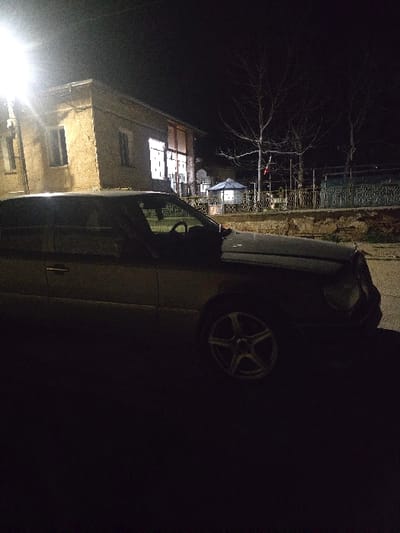 The scene depicts an outdoor nighttime environment in Gorna Mitropolia, Bulgaria. In the foreground, a dark-colored sedan, identified as consistent with a Mercedes-Benz W124 E-Class, is parked on a paved surface. The vehicle's driver's side and front are partially illuminated by an overhead streetlight, revealing multi-spoke alloy wheels. No occupants are visible within the vehicle.
To the left of the vehicle, a two-story building with a light-colored facade, possibly stucco or masonry, is visible. The ground floor of this building features large windows or glass doors that emit interior light. A bright street lamp on a pole stands to the far left, casting significant light on the building and the car's roof.
To the right of the parked vehicle, a low wall topped with a decorative metal fence separates the foreground from a slightly elevated area. Behind this wall and fence, a light-colored gazebo with a dark roof is situated. Several bare deciduous trees are visible in the background, along with a smaller structure featuring a blue roof further beyond. The environment is dark, with artificial light sources providing the primary illumination. No individuals are present in the visible area.