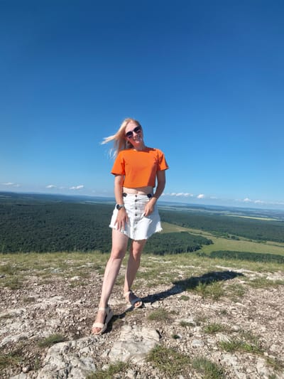 A full-body, slightly low-angle shot captures a blonde woman standing on a rocky, grassy cliff overlooking a vast, forested valley under a clear blue sky. She is positioned in the center-right of the frame, facing forward with her head tilted slightly to her left and a smile on her face. The woman is wearing an orange, cropped t-shirt, a white denim skirt, and light-colored wedge sandals. She has on dark sunglasses and a watch on her left wrist. Her blonde hair is blowing to her left in the wind. The background showcases a distant landscape of rolling hills covered in dense green forest, with a lighter green, grassy area visible in the foreground of the valley. The sky is a vibrant, cloudless blue. The ground in the foreground is composed of light-colored rocks and sparse green grass. The scene appears to be a scenic overlook, possibly in or near the city of Shikhan, Russia, given the expansive natural landscape.