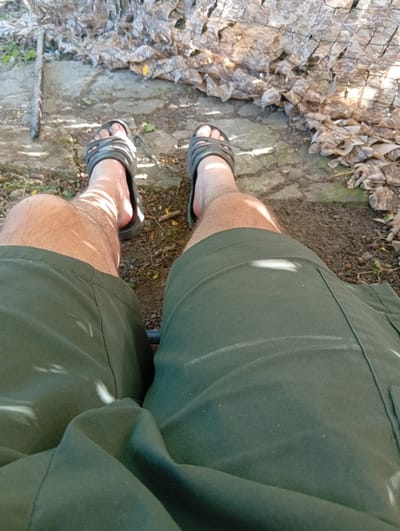 The image shows a close-up view of a person's legs and feet. The person is wearing dark green shorts and black sandals. They are sitting outdoors, with their feet resting on a paved surface and dirt. In the background, there is a camouflage net and some dry leaves. The lighting suggests it is daytime and there are shadows cast on the person's legs. The location is specified as Smyrnivka, Ukraine.