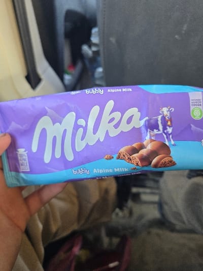 A human hand, visible on the lower left, presents a purple-foil-wrapped Milka "Bubbly Alpine Milk" chocolate bar. The wrapper features the "Milka" logo in white, a purple and white Alpine cow with a bell, and several aerated chocolate pieces. Text includes "bubbly," "Alpine Milk," "100% Alpine Milk chocolate," and a "Sustainably Sourced Cocoa" emblem. The background is indistinct, showing blurry light-colored and dark, textured surfaces consistent with a vehicle interior or cluttered room. The scene is located in the city of Opalchenets, Bulgaria.
