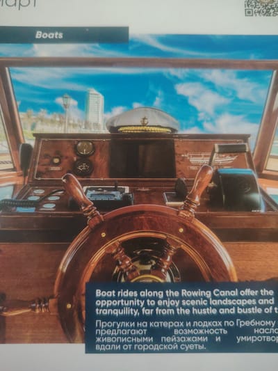 The image shows the interior of a boat, focusing on the captain's helm. A large wooden steering wheel is prominent in the foreground. Behind it, a polished wooden dashboard features various gauges, controls, and a radio. A white captain's hat with a gold braid band and an anchor emblem rests on the dashboard. The background depicts a bright blue sky with white clouds, and in the distance, a city skyline with buildings and palm trees. Text overlays on the image, partially in English and partially in Russian, advertise boat rides along the "Rowing Canal" offering "scenic landscapes and tranquility, far from the hustle and bustle of the city." The image does not contain any information about the city of Bahshitpa, Uzbekistan.
