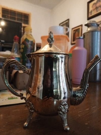 An ornate, silver-toned metal teapot or coffee pot is centrally positioned on a dark brown wooden table surface. The pot features a decorative handle on the left, an elaborate spout on the right, and three support feet with floral motifs. A conical lid with a finial, resembling a small pine cone or artichoke, crowns the vessel. Its highly reflective surface mirrors distorted elements of the surrounding room, including a partial reflection consistent with a person holding a camera device.

In the blurred background to the left, a dark electronic screen, possibly a television, is visible. Adjacent to it are a clear bottle with a red cap and a larger bottle with a yellow cap and a green/yellow label. A multi-paned window frame is discernible in the upper left background.

To the right in the background, an orange plastic container with a white lid, a white ceramic mug, and a light pink insulated water bottle are present. Further to the right, a tall, cylindrical silver metal container, consistent with a milk can or large thermos, is visible. Several framed pictures are displayed on a light-colored wall behind these objects.

No individuals are directly depicted within the scene. The objects are static, indicating no active interactions. The environment suggests an indoor domestic setting, likely a kitchen or dining area. The location is Longmiang, Laos.