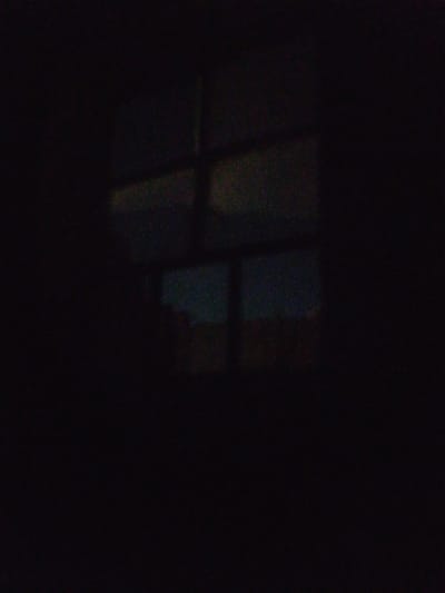 A nighttime photograph taken from inside a dark room, looking out at a window. The window has a grid of four panes and shows a faint reflection or scene from outside. The outside scene appears to be dimly lit, with blurry shapes and colors suggesting distant lights or structures. The overall image is dominated by darkness, with a slight grainy texture throughout. The location in Достук, Kyrgyzstan is not discernable from the visual information provided.
