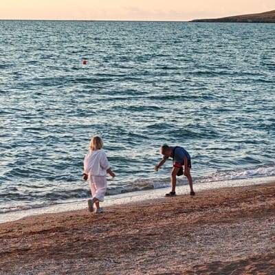 The image depicts a serene beach scene at dusk. Two individuals are present near the water's edge. A younger person, possibly a child, stands facing away from the camera, dressed in a light-colored outfit. To their right, a slightly older individual, dressed in a blue t-shirt and dark shorts, is bent over, seemingly examining something on the sand. The sea is calm with gentle waves lapping at the shore. In the distance, a small orange buoy floats on the water. The background shows a faint landmass and the horizon where the sky meets the sea, bathed in the soft hues of sunset. The beach itself is covered in sand and small shells.
