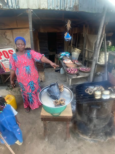 An adult female, wearing a blue headwrap and a red and teal patterned long dress, stands in front of a market stall in Idon Kasa, Nigeria. She points with her right hand towards a wooden display table laden with goods. On the table are multiple woven baskets filled with red onions, alongside clear plastic-wrapped packages of grains or seeds. A partially visible red and white "POS AVAILABLE" sign is affixed to a wooden structure behind her. To her immediate left, a yellow plastic jerrycan rests on the dirt ground, and a small child in a blue garment is partially visible in the bottom left. In the foreground, a green mechanical weighing scale with a silver tray holds a black plastic bag and several cylindrical, brownish items, likely smoked fish or snails. Adjacent to the scale, on the right, is a dark, cylindrical grill or smoker topped with a metal grate, on which numerous smoked items, including cylindrical bundles, are arranged. The stall itself features corrugated metal roofing and wooden support beams, with various dried items and a blue plastic bag hanging from the ceiling structure.