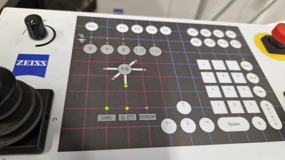 The image shows a close-up of a ZEISS brand control panel, likely for a scientific instrument. The panel is white with a black grid overlayed with pink and blue lines. There are numerous buttons and indicators on the panel.

Key elements visible include:
- A black rotary knob with a white indicator line on the upper left.
- The blue ZEISS logo on the upper left.
- A joystick-like control on the lower left.
- Circular buttons labeled ESC, F1 through F12.
- Circular buttons labeled X, Y, Z, and two with arrows indicating directional movement.
- A central circular graphic with "1-5" in the middle, connected by lines to several small yellow dots and one green illuminated dot.
- Several small circular indicator lights, some green and some yellow.
- Rectangular buttons labeled CNC, with a symbol that appears to be a crosshair within a circle, and ERROR.
- A numeric keypad with buttons labeled 0-9, minus, and period.
- Modifier buttons labeled Num, Shift, and Tab.
- Four circular directional buttons (up, down, left, right arrows).
- A large oval button labeled "Space".
- A rectangular button labeled "Return".
- A prominent red emergency stop button with a yellow surround on the upper right.

The lighting suggests an indoor environment, likely an industrial or laboratory setting. The image is taken from a slightly elevated angle, looking down at the control panel. There are no people visible in the image. The background is out of focus but appears to be shelves or equipment. The specific location in Ichenhausen, Germany, cannot be determined from the visual information in the image.