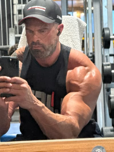 The image depicts a muscular man, likely Caucasian, taking a selfie in what appears to be a gym setting. He's wearing a black baseball cap with lettering visible, a dark-colored t-shirt, and holding a black smartphone. His right arm and shoulder are prominently displayed, showcasing well-defined musculature. A white towel is draped around his neck. The background reveals gym equipment, including weight plates and a metal rack. The environment suggests an indoor, artificially lit space. The location in the city of Jemelle, Belgium, is not discernible from the visual information provided.