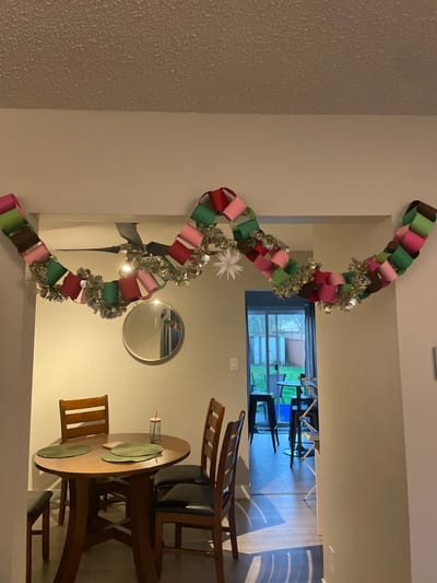 A brightly-lit dining area displays a Christmas-themed setting. A colorful paper chain garland with silver tinsel is draped across the wall, near a circular mirror. A round wooden dining table is set with green placemats. Wooden chairs surround the table. Through an open doorway, a glimpse of a room with stacked chairs and a window is visible.
