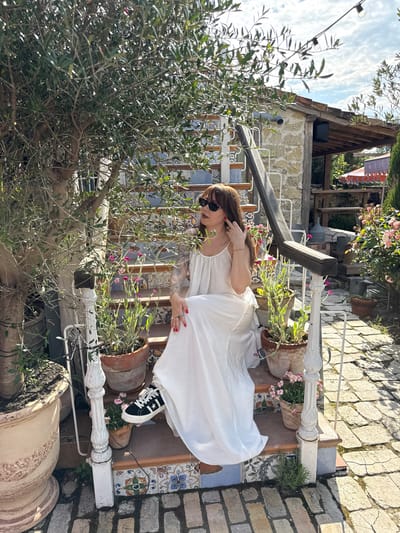 A woman with brown hair and sunglasses is sitting on outdoor stairs. She is wearing a white dress and black Converse sneakers. The stairs are decorated with colorful tiles and lined with potted plants. Behind her, there is a stone building and a large tree with olive branches. The ground is paved with cobblestones. The scene is set in a sunlit outdoor environment, with a blue sky and some clouds visible.