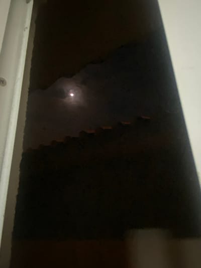 The image depicts an obscured night scene, possibly viewed through a window. A bright, circular light source, likely the moon, is visible through what appears to be an opening or break in a dark barrier or foliage. The foreground features a horizontal white structure, likely a window frame or sill. The overall environment is dark and shadowy. The location is purported to be La Aurora, Ecuador, but this cannot be verified from the image alone.