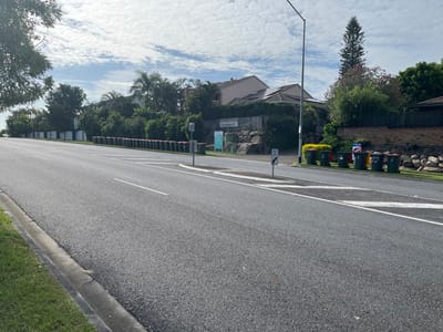 This bright, daytime image captures a quiet suburban street scene in Riverhills, Australia, under a partly cloudy sky. The asphalt road, appearing to run from the foreground to the background, is marked with clear white lines, including a solid curb line, a broken lane line, and a large directional arrow painted on its surface.

On the left side of the road, a row of household wheelie bins in assorted colors (green, yellow, red, and dark green) are positioned neatly at the curb, indicating they are ready for waste collection. Attached to one of these bins is a blue "FOR SALE" sign, with "MCDONALD" visible below the main text. Behind the bins, a stone retaining wall and a low brick fence separate the road from the properties. Further back, residential houses are partially obscured by abundant green trees, one of which is a tall, prominent conifer. A utility pole stands near the curb, and several smaller signs on posts are visible further down the street.

The right side of the road is edged by a concrete curb and a well-maintained strip of green grass. A circular speed limit sign, clearly displaying the number "70", is present on this side. Beyond the grassy verge, more landscaping features, including stone elements, and lush trees fill the background.

The image portrays a typical, well-kept suburban environment, with no people or specific activities taking place. The bright lighting suggests it is late morning or early afternoon.