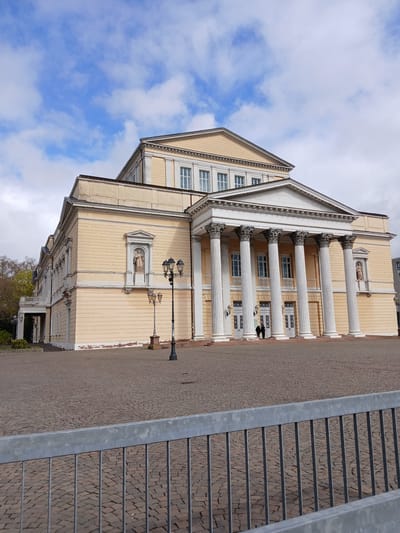 The image depicts the Staatstheater Darmstadt, located in Darmstadt, Germany. It is a neoclassical building with a pale yellow facade, featuring a prominent portico supported by white Corinthian columns. A statue is recessed into the wall on the left side of the structure. Two people can be seen near the entrance of the building. A vintage-style streetlight stands in front of the building on the paving stone street. A metal railing can be seen along a stairway, taking up the right side of the photo. The sky in the background has scattered white clouds.
