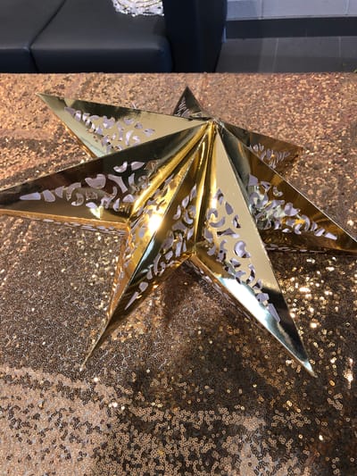 A close-up, eye-level shot displays a golden, three-dimensional star ornament lying on a table covered with a shimmering, rose gold sequined fabric. The star, which appears to be made of foil or laminated cardstock, has intricate cut-out patterns on its surface, revealing a white interior. The star is positioned slightly off-center, with its points extending towards the edges of the frame. The background is softly blurred, showing hints of dark furniture and a tiled wall, suggesting an indoor setting. The lighting is bright, creating reflections on the sequined fabric and the metallic surface of the star. No people are visible in the image. The location is not specified.