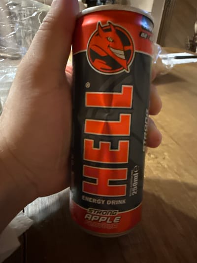 A hand is holding a red and black can of "Hell" energy drink. The can features a cartoon devil logo and text indicating "ENERGY DRINK" and "STRONG APPLE FLAVOUR". The background is blurred but appears to be a wooden surface, possibly a table, and some indistinct objects. There is no visible indication of the city of Goliam Dol, Bulgaria.