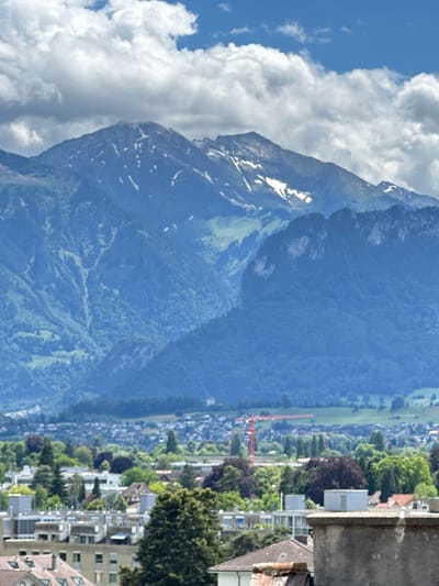 This image captures a panoramic view of Thun, Switzerland, with its iconic mountain range serving as a dramatic backdrop. The mountains, tinged with patches of snow on their upper reaches, dominate the upper half of the frame. Their slopes are a tapestry of verdant green, with visible valleys and ridges. Below the mountains, a dense urban landscape unfolds. Buildings of varying heights and architectural styles cluster together, interspersed with lush green trees. A prominent red construction crane is visible in the distance, suggesting ongoing development within the city. The sky above is a vibrant blue, dotted with fluffy white clouds. The foreground is occupied by rooftops and the upper facades of buildings, indicating the vantage point is from a higher elevation within the city.